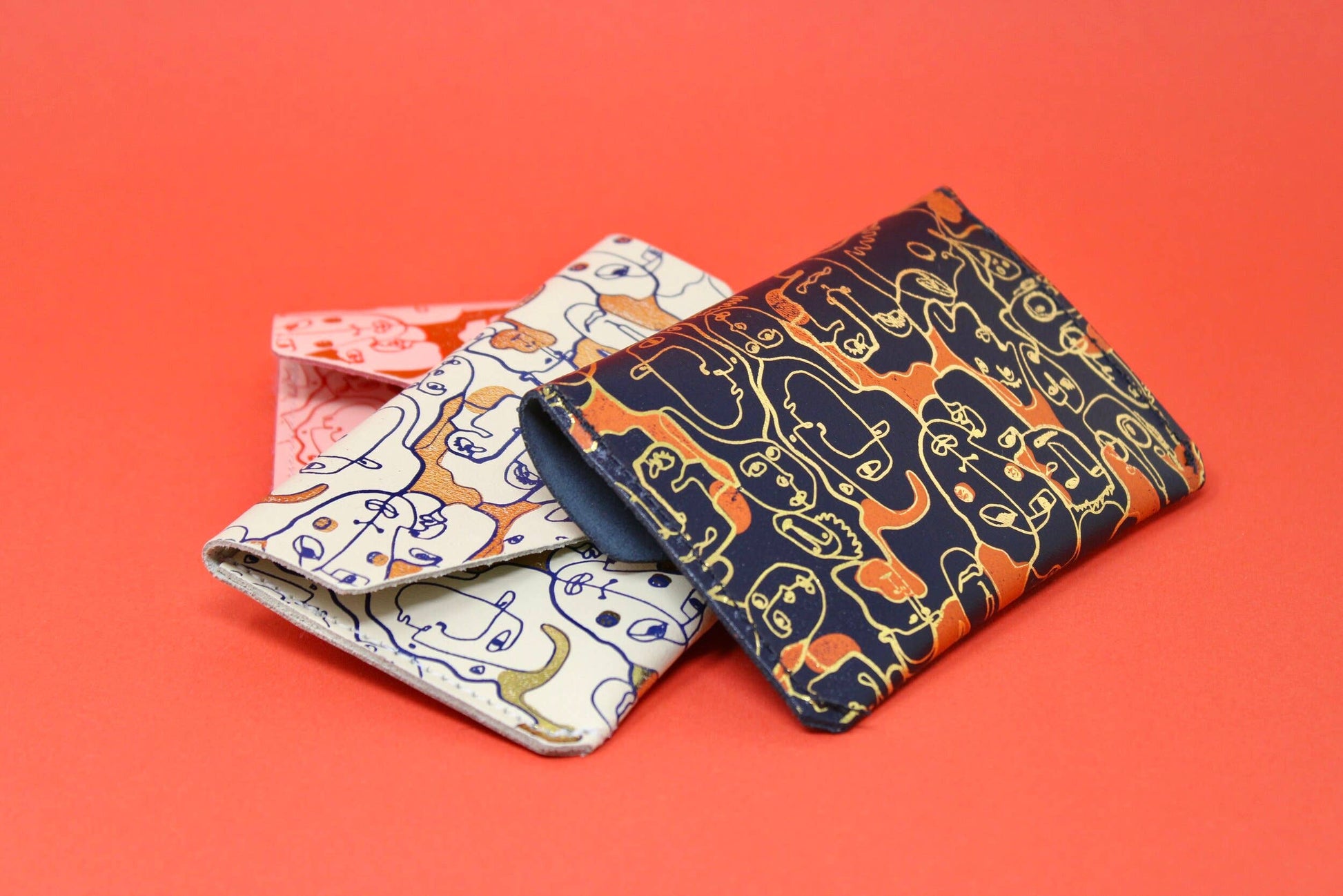 A small leather purse embossed with a wrap around figurative design. The simple line illustration features a collection of different characters. Purse is navy with gold embossed detail and patches of orange.