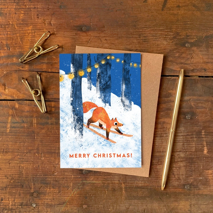 Illustrated Christmas greeting cards by Emily Nash Illustration. Snowy scenes of a fox skiing through a snowy wood with twinkling lights between the trees. With the words Merry Christmas!