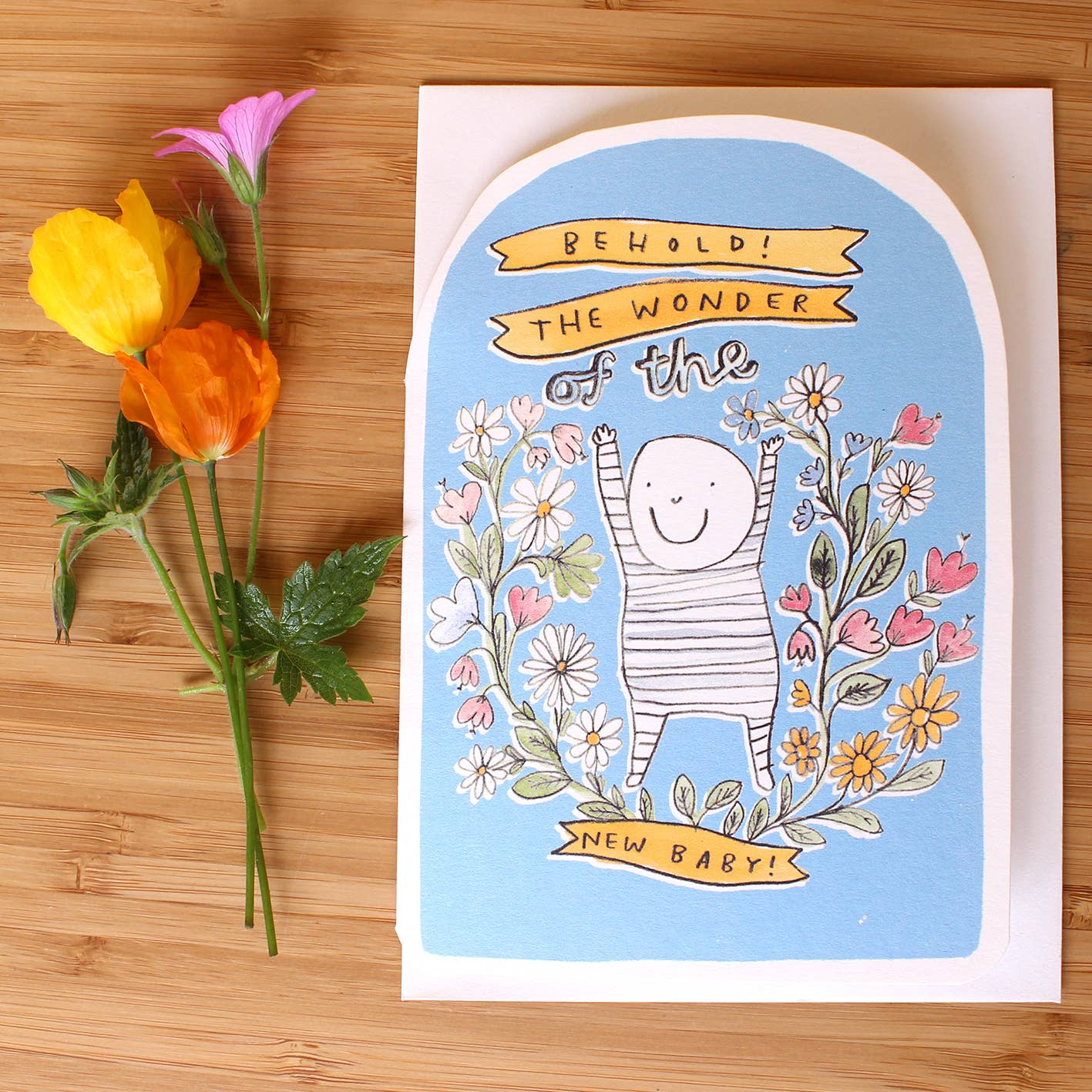 The words Behold! The wonder of the new baby! With an illustration in black and white of a smiling baby in a stripy babygrow. Surrounded by pink and yellow flowers, against a blue background. The card top is an oval.