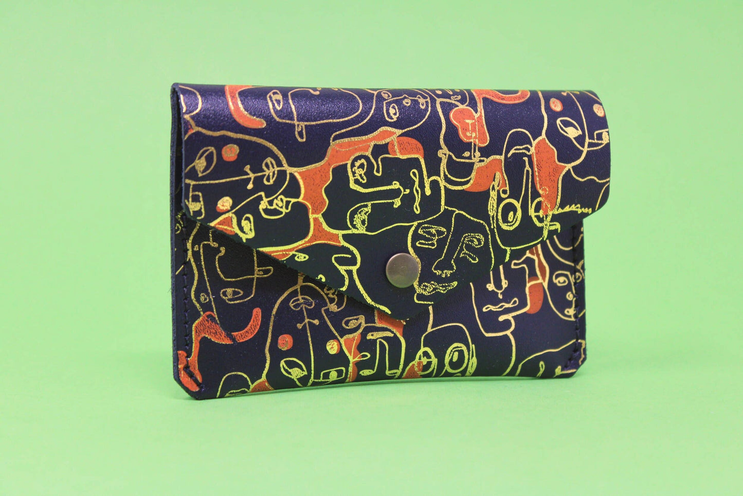 A small leather purse embossed with a wrap around figurative design. The simple line illustration features a collection of different characters. Purse is navy with gold embossed detail and patches of orange.