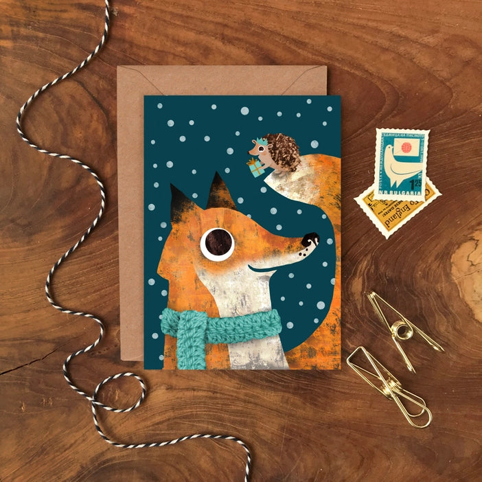 Illustrated Christmas greeting cards by Emily Nash Illustration. With the words Merry Christmas. featuring a hedgehog in a Christmas hat, presenting a gift to her friend a fox in a mustard cosy scarf. All the while the dark sky is filled with snow.