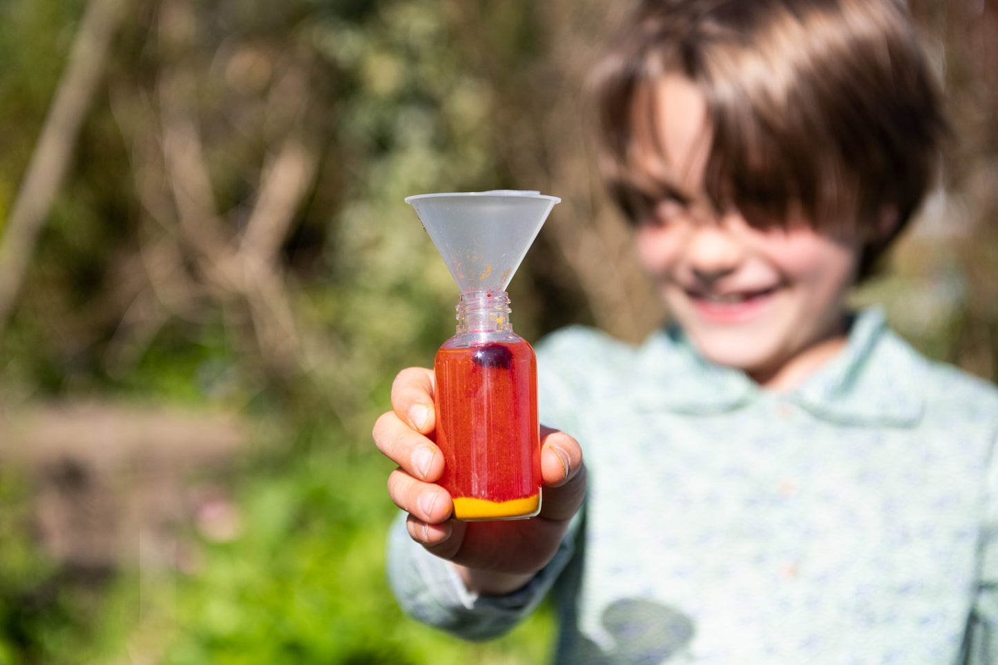 A child proudly holds up a bottle of red potion with a funnel sticking out of the top.