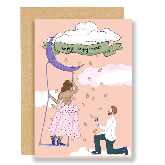 A dreamy card for a dreamy couple. This card is literally raining engagement rings from a cloud, all the while the woman stands on a swing hanging from the moon in a gorgeous backless pink dress with red lovebhearts all over it. As the man kneels down in his casual outfit and proposes. With the words at the top of the card, happy engagement.