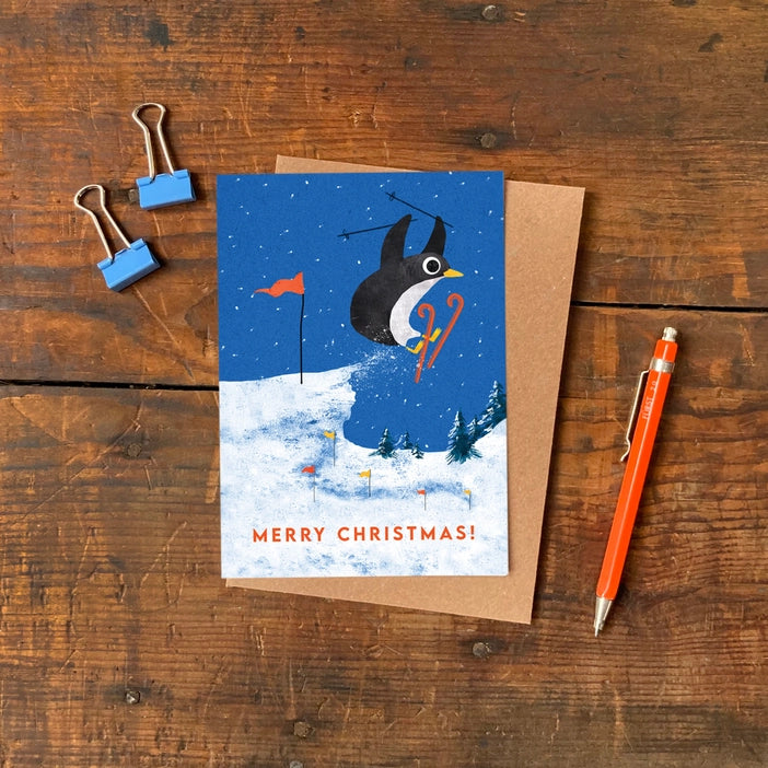 Illustrated Christmas greeting cards by Emily Nash Illustration. Snowy scenes of a penguin making a ski jump in a snowy scene. With the words Merry Christmas!