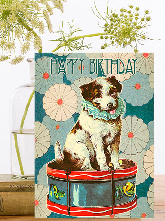 A whimsical birthday card featuring a charming pup in a fancy collar, perched on a drum, ready to serenade you with a catchy beat. With the words Happy Birthday at the top of the card.